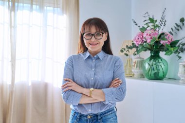 Oturma odasında poz veren orta yaşlı bir kadının portresi, kendinden emin 45 yaşında kadın kameraya bakıyor, kolları bağlı duruyor.