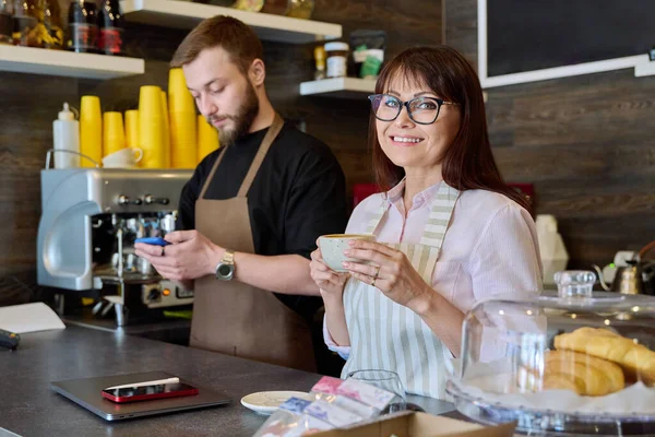 Team, partners, young male and mature woman talking working using phone standing behind bar in coffee shop. Team, small business, work, staff, cafe cafeteria restaurant, entrepreneurship concept