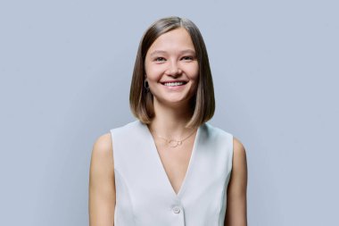 Gri stüdyo arka planında zarif bir iş kadınının portresi. Beyaz yelekli, doğal makyajlı, gülümseyen kadın kameraya bakıyor. Gençlik, güzellik, iş, eğitim, insan kavramı