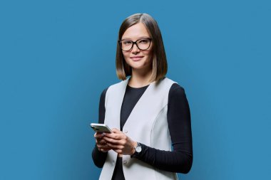 Mavi stüdyo geçmişinde akıllı telefonu olan kendine güvenen genç bir kadın. Gözlüklü gülümseyen kadın kameraya bakıyor. İş için mobil uygulamalar, eğitim, eğlence, teknoloji, insanlar.