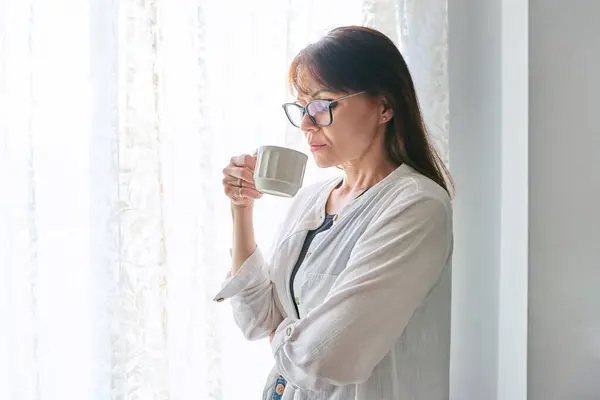 Serious middle-aged woman at home looking out window, with cup in her hands. Calmness, relaxation, loneliness, mature age people concept