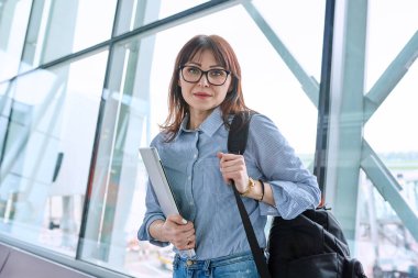 Sırt çantalı orta yaşlı bir kadın havaalanı terminalinde uçak bileti bekliyor, dizüstü bilgisayarını tutuyor, kameraya bakıyor. Seyahat, iş gezisi, yolcu taşımacılığı konsepti