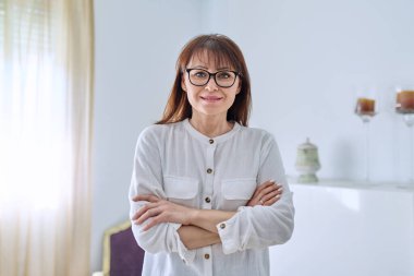 Oturma odasında poz veren orta yaşlı kadın portresi, kameraya bakarak kollarını kavuşturan 45 yaşında kendinden emin bir kadın. Yaşam tarzı, ev, olgun insanlar konsepti