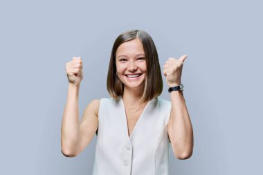 Neşeli, neşeli, mutlu, yumruklarını sıkan genç bir kadın gri stüdyo arka planında el ele tutuşuyor, evet jest. Mutluluk, neşe, sürpriz, iyi haber, zafer, başarı, kazanan, gençlik kavramı