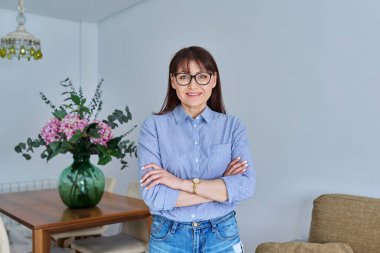 Oturma odasında poz veren orta yaşlı bir kadının portresi, kendinden emin 45 yaşında kadın kameraya bakıyor, kolları bağlı duruyor. Yaşam tarzı, ev, olgun insanlar konsepti