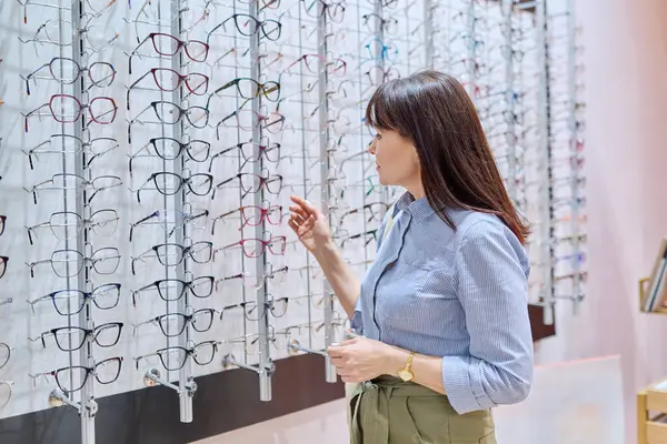 Middle-aged woman in opticians store buying, choosing glasses frames, near display case with vision eyeglasses spectacles. Optics, ophthalmology, age vision problems