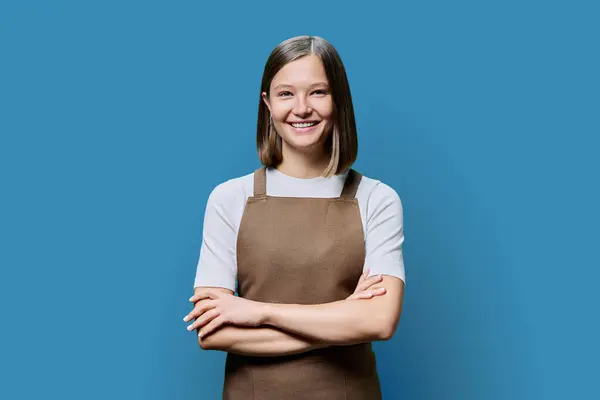 Portrait of young smiling confident woman in apron on blue studio background. Successful positive female with crossed arms. Worker, startup, small business, service sector, staff, youth concept