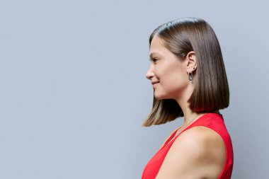 Gri stüdyo arka planında saç stili modaya uygun doğal makyajlı, kırmızı elbiseli genç bir kadının profil görüntüsü, reklam metni için alan kopyalama. Güzellik, moda, makyaj, yüz kılları.