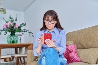 Akıllı telefonlu orta yaşlı bir kadın, evinde oturma odasında, 40 'lı yaşlarda cep telefonu kullanan gözlüklü bir kadın. Yaşam tarzı, iletişim, rahatlık, serbest çalışma, olgun insanlar konsepti