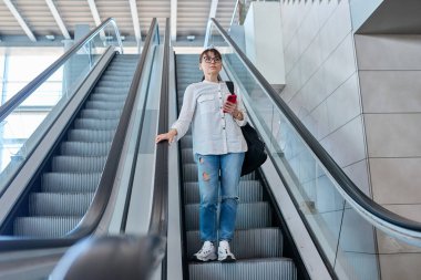 Sırt çantalı orta yaşlı bir kadın yürüyen merdivende, modern istasyon binasında, havaalanında günlük kot pantolon giyiyor.