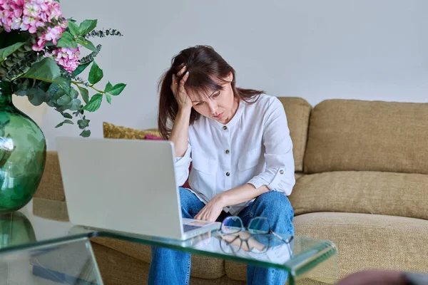 疲れた中年の女性は頭痛に苦しみ リビングルームの自宅でラップトップコンピュータでソファーに座っていました ホームオフィスで働く40代女性 疲れた 片頭痛 精神的ストレス — ストック写真