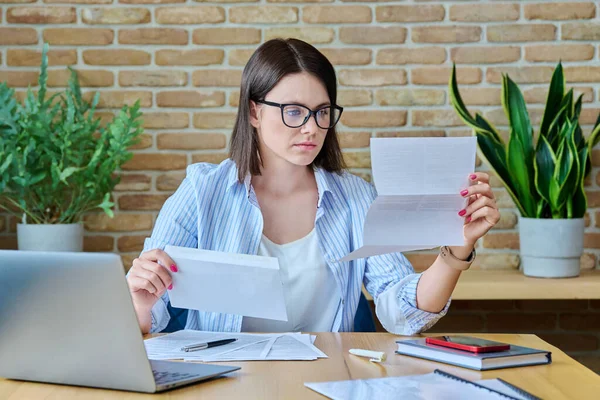 Serious young business woman reading important official letter sitting at desk in office. Business work banking services taxes, contract statement agreement investment deal, paperwork correspondence