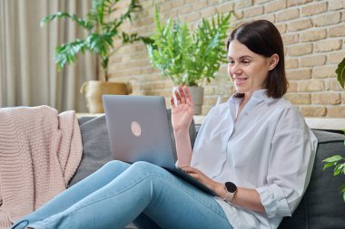 Oturma odasındaki kanepede oturan rahat genç bir kadın görüntülü konuşma için dizüstü bilgisayar kullanıyor. Tatiller, eğlence, teknoloji, konfor, yaşam tarzı, gençlik kavramı