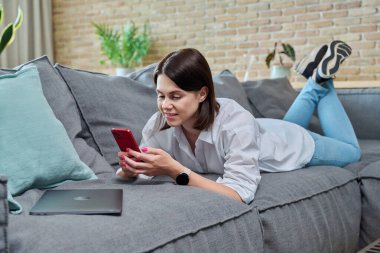 Genç, mutlu, akıllı telefon kullanan, evdeki kanepede uzanan bir kadın. İnternet çevrimiçi hizmetler, iletişim, mobil uygulamalar, eğlence eğitimi için teknolojiler iş yaşam tarzı