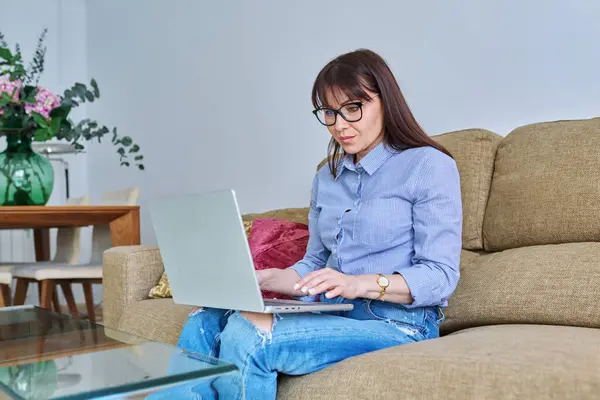 リビングルームのソファーに座って ノートパソコンのキーボードをタイプする中年女性 レジャーワークコミュニケーションのためのコンピュータを使用して深刻な成熟した女性 ソーシャルメディアブログ フリーランスのインターネット技術の人々 — ストック写真