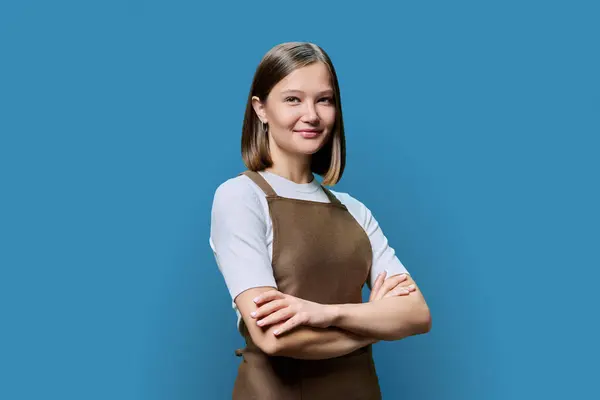 Portrait of young smiling confident woman in apron on blue studio background. Successful positive female with crossed arms. Worker, startup, small business, service sector, staff, youth concept