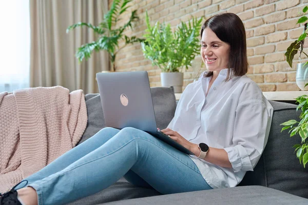 Young relaxed woman sitting on sofa at home in living room, using laptop for work, freelancing, learning, blogging, communicating, typing on keyboard
