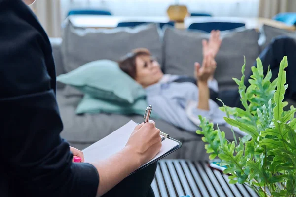 stock image Close-up of female psychologists hands with clipboard making notes, in individual therapy session. Woman patient lying in couch out of focus. Psychology psychotherapy treatment mental health concept