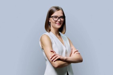 Gri stüdyo arka planında kollarını kavuşturmuş, gözlüklü, gülümseyen genç bir kadın. Pozitif iş, kendinden emin başarılı bir kadın kameraya katlanmış elleriyle bakıyor. İş, iş, eğitim, insanlar