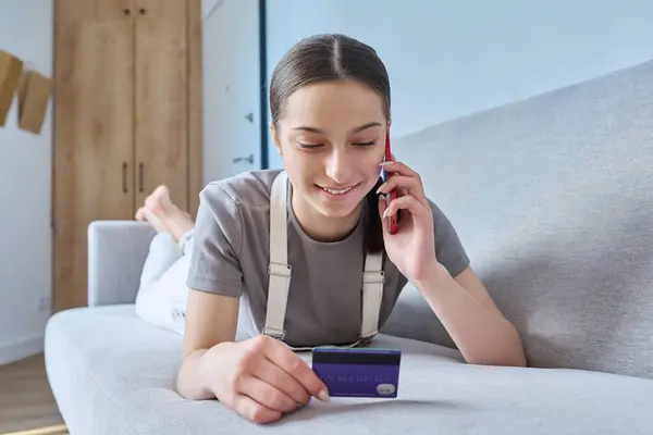 Young teenage student girl with credit bank card talking on cell phone lying on sofa at home. Online payment for goods and services, shopping, credit cards for teenagers concept