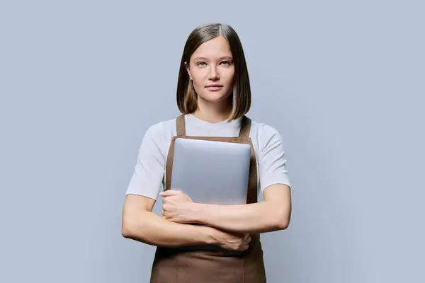 Portrait of young smiling confident woman in an apron holding laptop in hands, looking at camera on gray studio background. Worker, startup, small business, job, service sector, staff, youth concept
