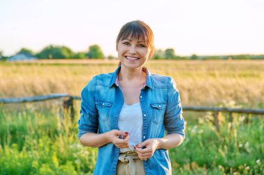 Gülümseyen orta yaşlı kadın portresi kameraya bakıyor, kırsal kesimde gün batımı doğa arka planında. Kendinden emin kadın çiftçi, çapraz kollu işçi.