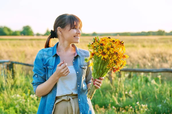 Profile view happy smiling joyful middle aged woman with bouquet of yellow flowers looking at setting sun wildlife background. Summer vacation weekend in nature, sunset, age beauty 45 years old female