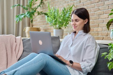 Oturma odasındaki kanepede rahat bir kadın oturuyor, iş için dizüstü bilgisayar kullanıyor, serbest çalışıyor, öğreniyor, blog yazıyor, iletişim kuruyor, klavyede daktilo yazıyor.