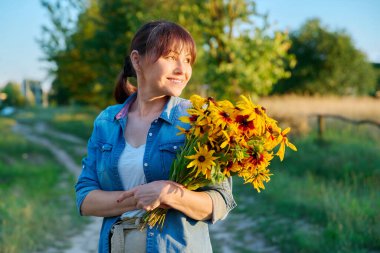 Profil görüntüsü mutlu gülümseyen orta yaşlı kadın bir buket sarı çiçekle vahşi yaşam arka planına bakıyor. Yaz tatili hafta sonu doğada, gün batımı, yaş güzelliği 45 yaşında kadın.