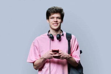 Kulaklıklı, akıllı telefonlu, gri arka planlı, yakışıklı genç bir erkek. Genç bir üniversite öğrencisi sırt çantasıyla kameraya bakıyor. İnternet teknolojisi, mobil uygulama hizmetleri