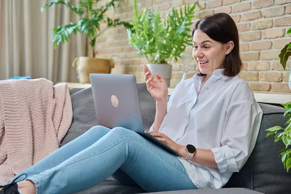 Oturma odasındaki kanepede oturan rahat genç bir kadın görüntülü konuşma için dizüstü bilgisayar kullanıyor. Tatiller, eğlence, teknoloji, konfor, yaşam tarzı, gençlik kavramı