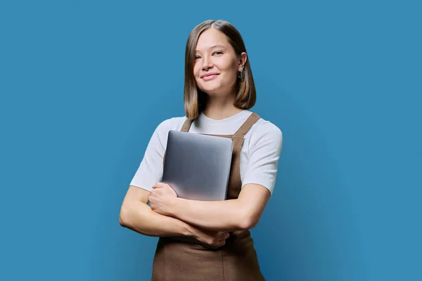 Portrait of young smiling confident woman in an apron holding laptop in hands, looking at camera on blue studio background. Worker, startup, small business, job, service sector, staff, youth concept