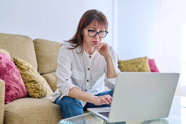 ラップトップコンピュータで働く真剣な中年の女性は リビングルームのソファーの自宅のオフィスに座っています リモートビジネス フリーランス 起業家精神 成熟したビジネス人々の概念 — ストック写真