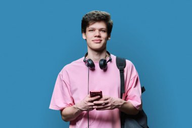Kulaklıklı, akıllı telefonlu, mavi arka planlı, yakışıklı genç bir erkek. Genç bir üniversite öğrencisi sırt çantasıyla kameraya bakıyor. İnternet teknolojisi, mobil uygulama hizmetleri