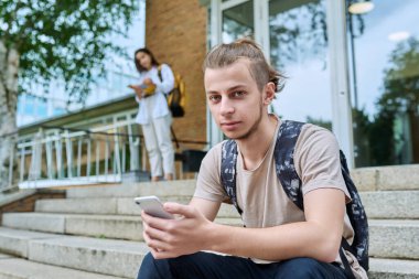 Hippi, 18-19 yaşlarında, sırt çantalı, akıllı telefonlu, açık hava eğitim binasının merdivenlerine oturmuş gülümseyen bir genç. Gençlik, eğitim, yaşam tarzı, teknoloji