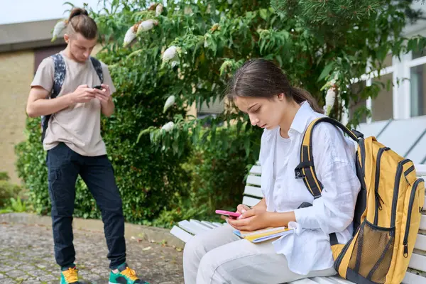 Teenage high school students, guy and girl, with backpacks near academic building using smartphones. Adolescence, youth, education, lifestyle, technology concept