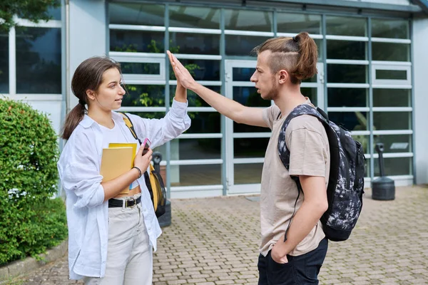 Meeting of two teenage students, guy and girl, outdoors near educational building, hand greeting. Friendship, communication, education, high school college concept
