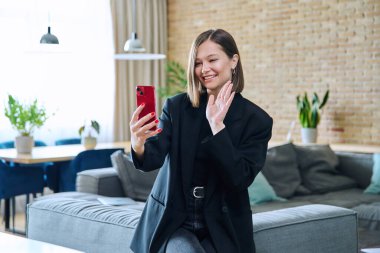 Oturma odasındaki koltukta oturan neşeli, genç bir kadın akıllı telefon kullanarak görüntülü konuşma yapıyor. İletişim, eğlence, ev, yaşam tarzı, gençlik, teknoloji kavramı