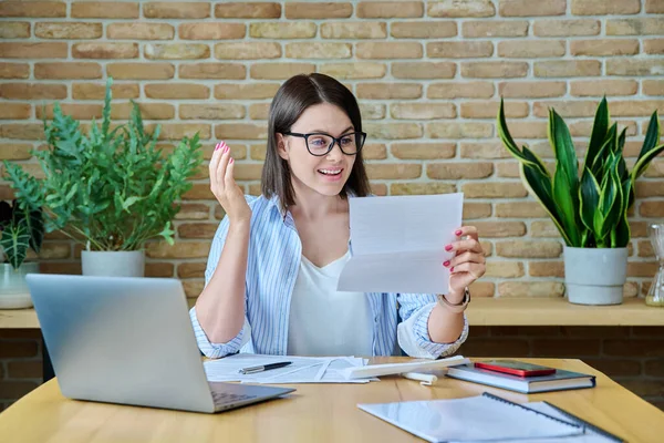 Young business woman opening paper envelope reading important letter, emotionally rejoicing at good news, sitting in office. Happiness joy winner, success in business, bank correspondence, work, study