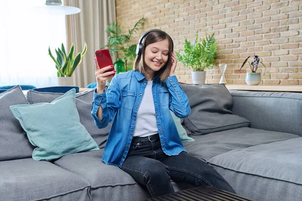 Young happy attractive cheerful woman in headphones listening to music, sitting on sofa at home, in living room, singing along with her eyes closed. Rest, lifestyle, fun, joy, good mood, youth concept