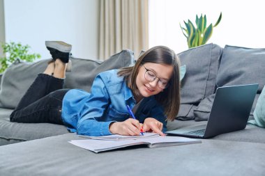 Evde ders çalışan genç bir üniversite öğrencisi dizüstü bilgisayar kullanarak kanepede uzanıyor. Öğrenim için eğitim bilgisi gençlik mobil uygulama İnternet çevrimiçi hizmet