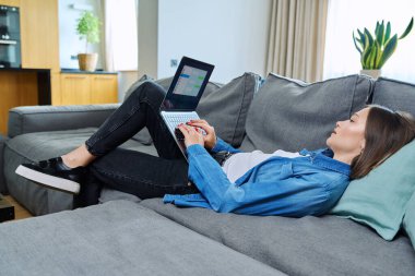 Oturma odasındaki kanepede uzanmış genç bir kadın bilgisayarı internet iletişimi için kullanıyor arkadaşlarıyla, iş arkadaşlarıyla, ailesiyle sohbet ediyor. İnsanlar, teknoloji, yaşam tarzı konsepti