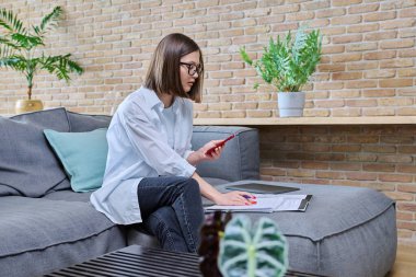 Genç kız üniversite öğrencisi evde oturuyor, kanepeye oturuyor, oturma odasında, defterde akıllı telefon yazıları kullanıyor. Eğitim bilgisi gençlik mobil uygulamaları İnternet çevrimiçi öğrenim hizmetleri