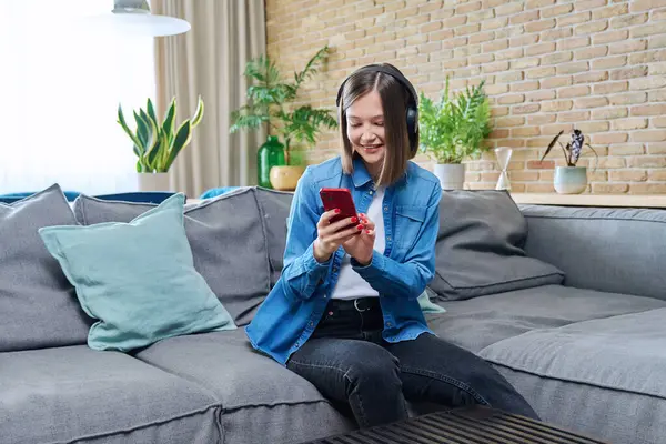 Young woman sitting on sofa in living room wearing headphones using smartphone, happy relaxing female listening music audio book learning languages chatting with friends, 20s student studying online