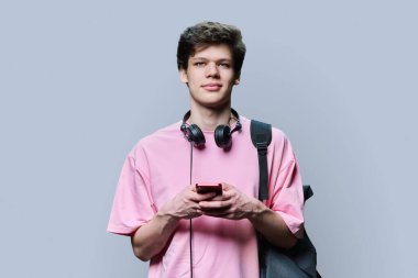Kulaklıklı, akıllı telefonlu, gri arka planlı, yakışıklı genç bir erkek. Genç bir üniversite öğrencisi sırt çantasıyla kameraya bakıyor. İnternet teknolojisi, mobil uygulama hizmetleri