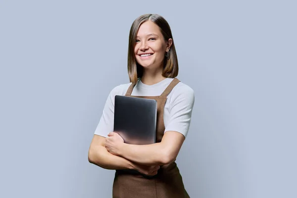 Portrait of young smiling confident woman in an apron holding laptop in hands, looking at camera on gray studio background. Worker, startup, small business, job, service sector, staff, youth concept