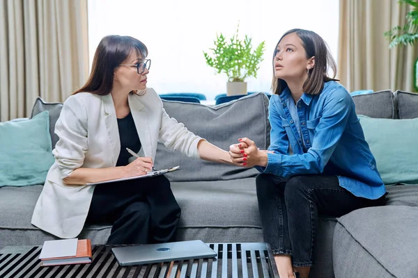 Sad young 20s woman in meeting with female psychologist therapist counselor sitting on couch in office. Psychology, therapy, psychotherapy, treatment, mental health concept