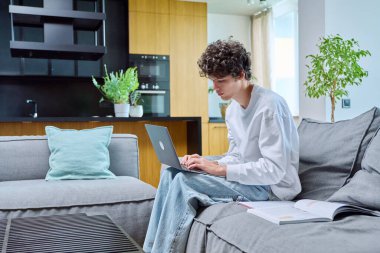 Yakışıklı bir üniversite öğrencisi, evde çalışıyor, kanepede oturuyor ve dizüstü bilgisayarını daktilo ediyor. Genç erkek, 18, 19 yaşında, oturma odasının içinde kıvırcık saçlı. Eğitim bilgileri çevrimiçi internet teknolojisi