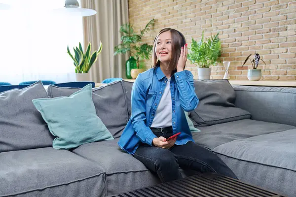 Young happy attractive cheerful woman in headphones listening to music, sitting on sofa at home, in living room. Rest, lifestyle, fun, joy, good mood, youth concept