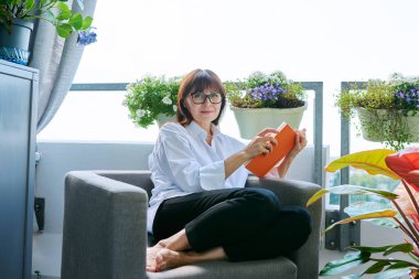 Orta yaşlı, rahat, mutlu bir kadın kitap okuyor, evdeki balkon terasında sandalyede oturuyor, kameraya bakarak gülümsüyor. Dinlenme, rahatlama, sakinlik, yaşam tarzı, rahatlık, okuma, olgun insanlar.